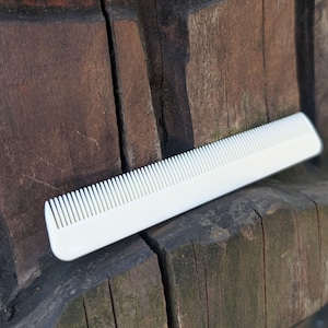 May include: A white hair comb with a straight spine and a row of fine, evenly spaced teeth. The comb features a smooth, matte finish and a rectangular shape with slightly rounded corners on one end, presenting a clean and functional design.