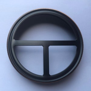 Puede incluir: Un anillo de metal negro con un borde decorativo de cobre. El anillo tiene un dise&ntilde;o en forma de T en el centro.