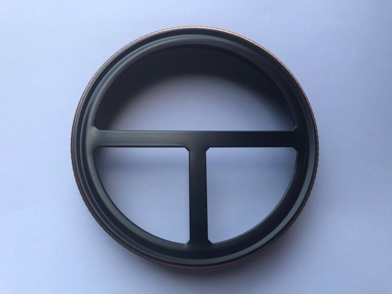 Puede incluir: Un anillo de metal negro con un borde decorativo de cobre. El anillo tiene un dise&ntilde;o en forma de T en el centro.