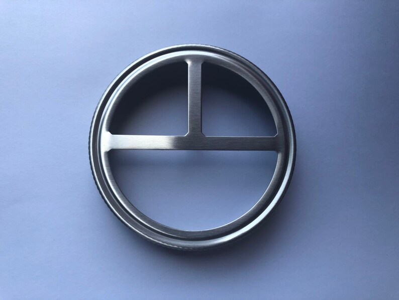 Puede incluir: Un anillo de metal plateado con un dise&ntilde;o circular que presenta tres barras que se cruzan. El anillo est&aacute; dise&ntilde;ado para encajar sobre una tapa de frasco.