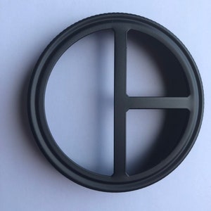 Puede incluir: Anillo de metal negro con cuatro recortes, formando una cruz. El anillo es circular y tiene un acabado liso y pulido.