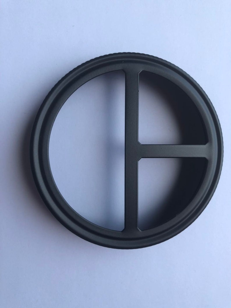 Puede incluir: Anillo de metal negro con cuatro recortes, formando una cruz. El anillo es circular y tiene un acabado liso y pulido.