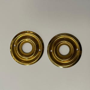 May include: Two antique brass door rosettes with concentric circular ridges and a central hole. The rosettes are a warm gold colour and have two small holes for mounting. These decorative hardware pieces are likely for a vintage door.