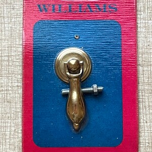 May include: A gold-toned cabinet pull with a round base and a long, slender handle. The pull is packaged on a blue and red card with the word "WILLIAMS" printed on it.