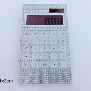 以下が含まれることがあります： 銀色のラインストーンのデザインが施された白い電卓。電卓は赤いディスプレイと、数字、演算子、関数を含む標準的なボタンレイアウトを備えています。電卓のサイズは19cm x 11cm x 3cmです。