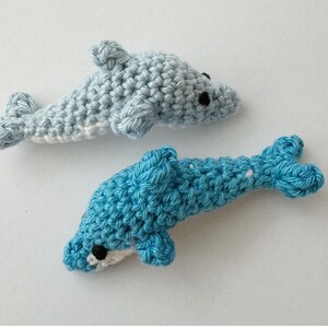 May include: Two crocheted dolphin toys. One is light blue, the other is a darker blue. Both have black eyes and white bellies. The dolphins are handmade and appear soft and cuddly. They are lying on a white surface.