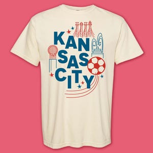 May include: Cream-colored t-shirt with a blue and red graphic that says "KANSAS CITY." The design includes illustrations of a soccer ball, tennis racket, and other city landmarks. The shirt has a crew neck and short sleeves.