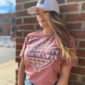 May include: A woman wearing a dusty rose t-shirt with a white graphic of a globe with the text "Kansas City I'm so in love" and a white baseball cap with a black logo.