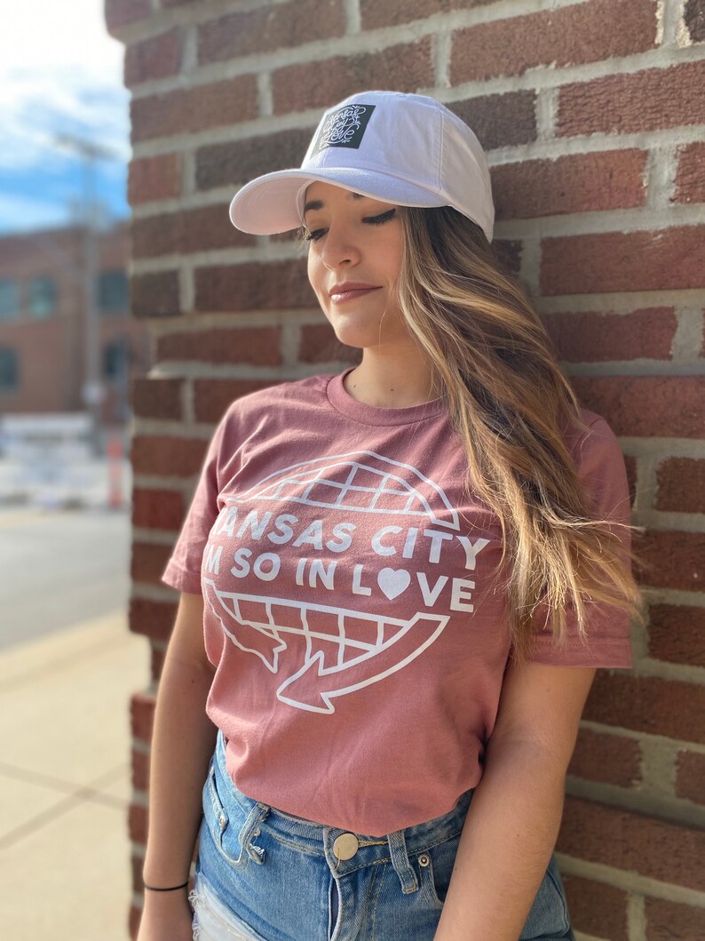 May include: A woman wearing a dusty rose t-shirt with a white graphic of a globe with the text "Kansas City I'm so in love" and a white baseball cap with a black logo.