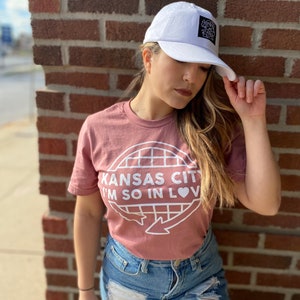 May include: A woman wearing a pink t-shirt with the text "Kansas City I'm so in love" and a white baseball cap with a black patch. She is wearing blue jeans with rips and holes.