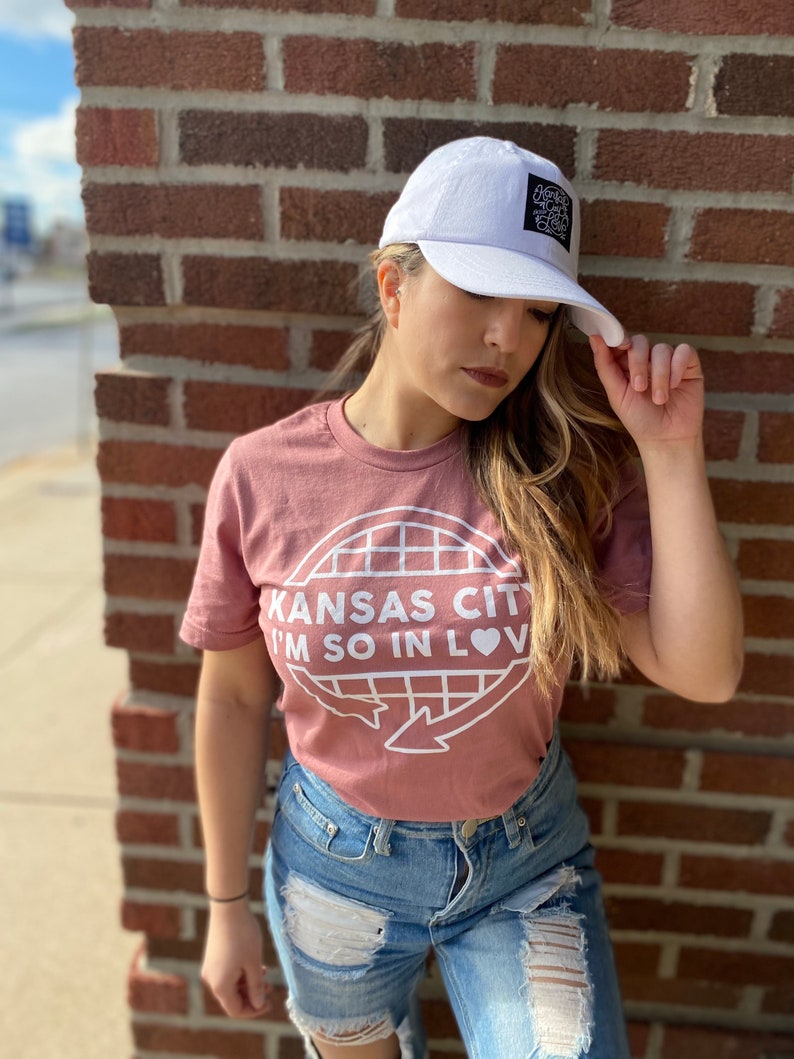 May include: A woman wearing a pink t-shirt with the text "Kansas City I'm so in love" and a white baseball cap with a black patch. She is wearing blue jeans with rips and holes.