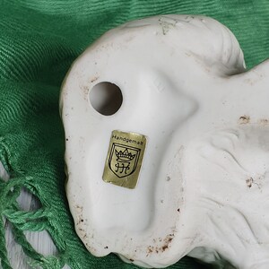 May include: White ceramic figurine of a dog with a gold label on the bottom that reads "Handgemalt" with a crown and the letters "JFC".