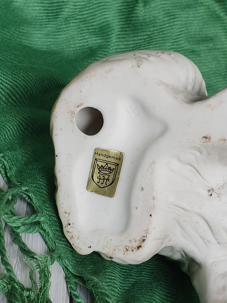 May include: White ceramic figurine of a dog with a gold label on the bottom that reads "Handgemalt" with a crown and the letters "JFC".