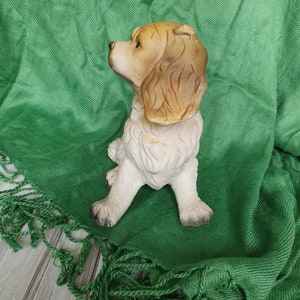 May include: A ceramic figurine of a brown and white dog sitting on a green fringed blanket. The dog is looking to the left and has floppy ears.