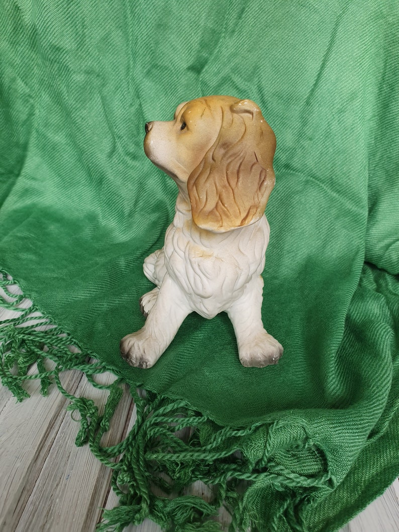 May include: A ceramic figurine of a brown and white dog sitting on a green fringed blanket. The dog is looking to the left and has floppy ears.