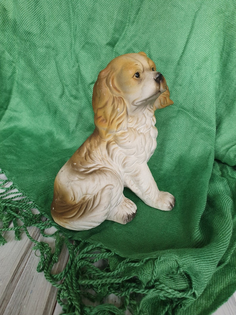 May include: A ceramic figurine of a brown and white dog with floppy ears. The dog is sitting on a green fabric background.