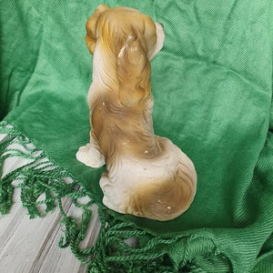 May include: A ceramic figurine of a brown and white dog sitting with its head turned to the side. The dog is on a green fringed fabric.