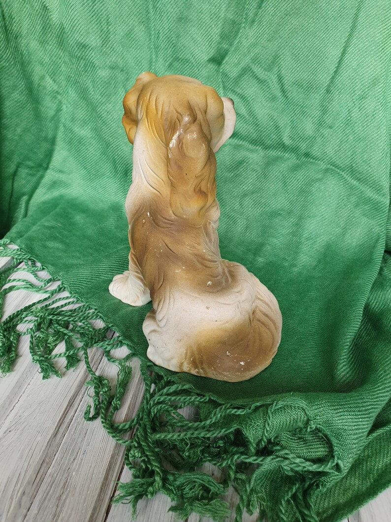 May include: A ceramic figurine of a brown and white dog sitting with its head turned to the side. The dog is on a green fringed fabric.