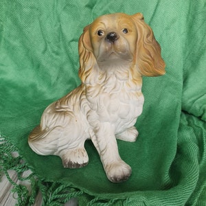 May include: A white and brown ceramic figurine of a dog sitting on a green fringed fabric. The dog has floppy ears and a brown nose.