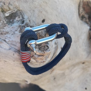 May include: A navy blue rope bracelet with a silver metal clasp. The rope has a red and gray stripe.