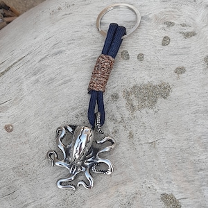 Könnte beinhalten: Ein silberner Oktopus-Schlüsselanhänger mit einem blauen und braunen Paracord-Lanyard. Der Schlüsselanhänger ist an einem silbernen Schlüsselring befestigt.