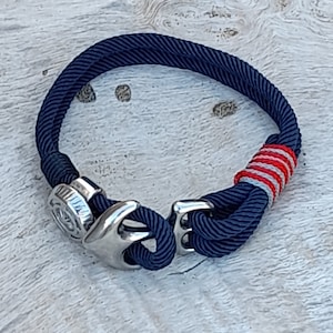 Puede incluir: Un brazalete de cuerda azul marino con un cierre de ancla de plata y un acento a rayas rojas y blancas.