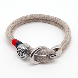 Op de afbeelding: Een beige touw armband met een zilveren anker sluiting en een kompas bedel. Het touw is gevlochten en heeft rode en blauwe accenten.