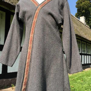 Könnte beinhalten: Graues Tunika-Kleid mit langen Ärmeln und Zierband. Das Band ist beige mit einem sich wiederholenden roten geometrischen Muster. Das Kleid wird auf einer Schaufensterpuppe im Freien mit einem reetgedeckten Gebäude im Hintergrund präsentiert.