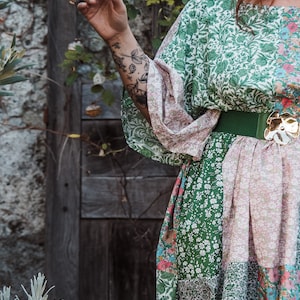 Può includere: Una donna indossa un abito a fiori verde e rosa con una cintura verde e una fibbia dorata. L'abito ha un design patchwork ed è realizzato in un tessuto leggero.