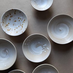 Könnte beinhalten: Sechs Keramikschalen mit verschiedenen Glasurmustern. Die Schalen sind weiß mit braunen und weißen Wirbeln und Flecken.