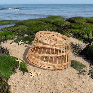 May include: A round, woven wicker lobster trap with a spiraled top design and open-slat sides. The light brown, natural-coloured basket is positioned on a sandy surface alongside four small, light-coloured starfish arranged near the structure.