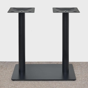 May include: Black metal table base with two vertical supports and a rectangular base. Each support has a square top with screw holes. The base is designed for stability and supports a tabletop. The supports are approximately 71 cm tall.