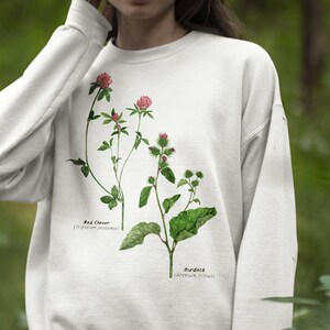 May include: White sweatshirt with a botanical illustration of red clover and burdock plants. The plants are labeled with their scientific names.