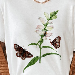 May include: White t-shirt with a floral design featuring a white flower with green leaves and two black and red butterflies.