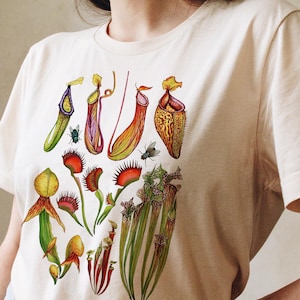 Peut inclure: T-shirt crème avec une illustration botanique colorée de diverses plantes carnivores, dont des plantes carnivores et des dionées attrape-mouches. Le motif présente des dessins détaillés des plantes avec des insectes, dans des tons de vert, de rouge, de jaune et d'orange.