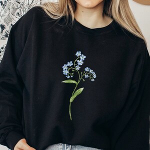 May include: Black sweatshirt featuring a watercolor illustration of a forget-me-not flower. The floral design includes light blue blossoms and green leaves on a stem. The sweatshirt is a crew neck style.