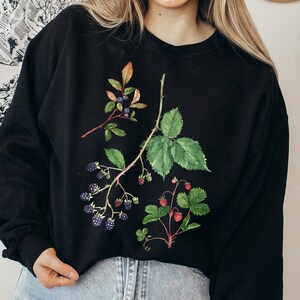 May include: Black sweatshirt featuring watercolor illustrations of berries and leaves. The design includes branches of blueberries, blackberries, and strawberries with green leaves and red berries. The sweatshirt is a crew neck style.