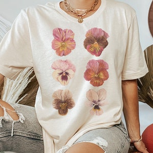 May include: A beige t-shirt with a graphic print of six pink, brown, and yellow pansies.