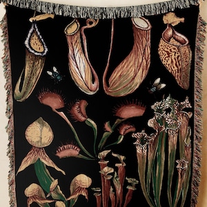 May include: Black throw blanket with botanical illustrations of carnivorous plants, such as pitcher plants and Venus flytraps. The artwork uses brown, green, and red tones, and has a fringed border. The blanket is displayed against a neutral background.