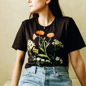 May include: A black t-shirt with a floral print featuring orange, yellow, and white flowers. The shirt is worn by a person with long dark hair and blue denim shorts.