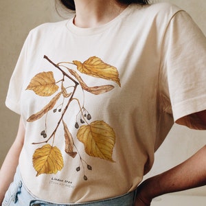 May include: Cream-colored t-shirt featuring a watercolor illustration of a linden tree branch with golden leaves and seed pods. The text "Linden tree (Tilia cordata)" is printed below the illustration. The shirt is paired with blue jeans.
