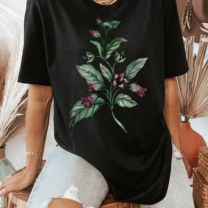 May include: Black t-shirt with a floral print of a green plant with purple flowers. The plant has dark green leaves and the flowers are a deep purple color.