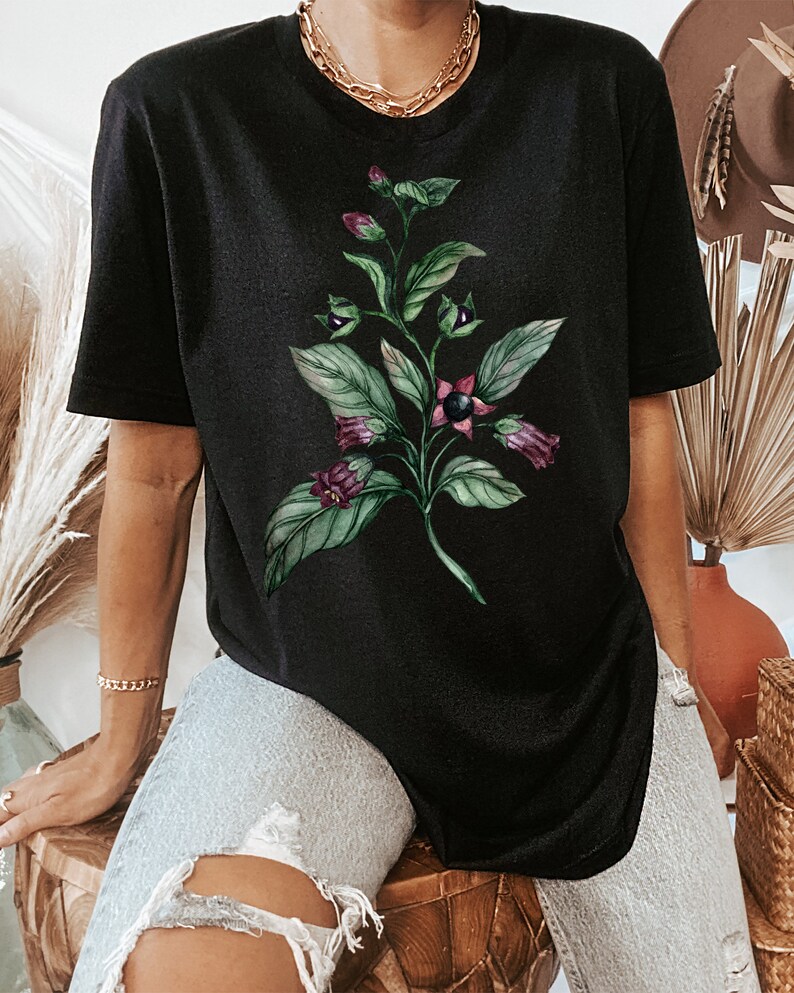 May include: Black t-shirt with a floral print of a green plant with purple flowers. The plant has dark green leaves and the flowers are a deep purple color.