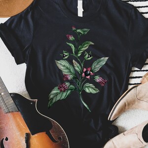 以下が含まれることがあります： 黒いTシャツに、水彩で描かれた緑色の植物と紫色の花が描かれています。植物には黒い実がなっていて、黒い背景に描かれています。