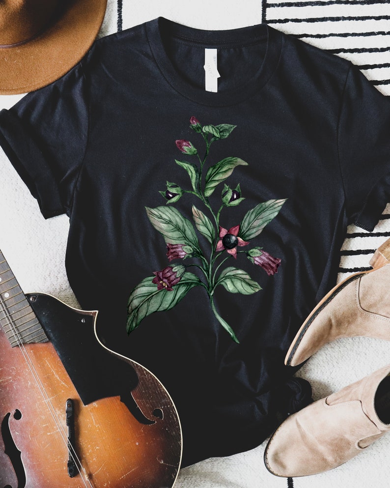 以下が含まれることがあります： 黒いTシャツに、水彩で描かれた緑色の植物と紫色の花が描かれています。植物には黒い実がなっていて、黒い背景に描かれています。