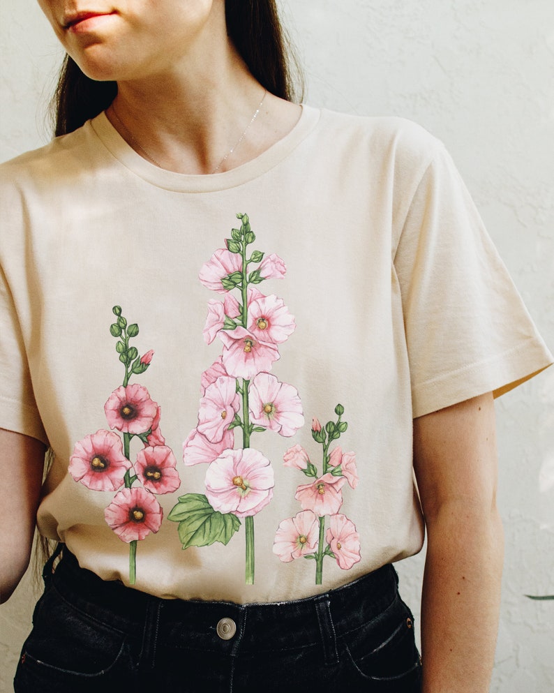 May include: A beige t-shirt with a floral print of pink hollyhock flowers on the front.