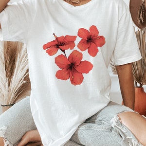 Może przedstawiać: Biała, oversizowa koszulka z nadrukiem graficznym trzech czerwonych kwiat&oacute;w hibiskusa.