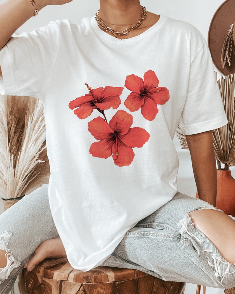 Może przedstawiać: Biała, oversizowa koszulka z nadrukiem graficznym trzech czerwonych kwiat&oacute;w hibiskusa.