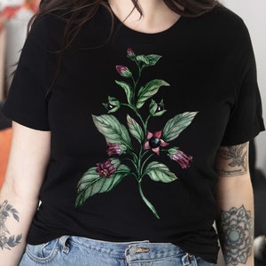 以下が含まれることがあります： 紫色の花が咲いた緑の植物が描かれた黒いTシャツ。植物は黒い実と緑の葉をつけています。水彩画で描かれています。