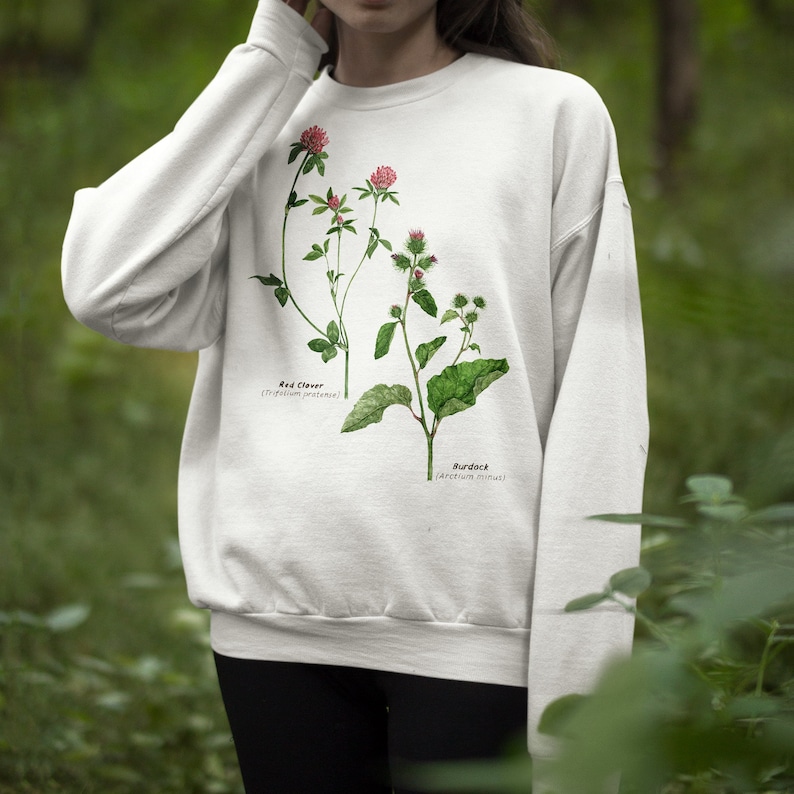 May include: White sweatshirt with a botanical illustration of red clover and burdock plants. The plants are labeled with their scientific names.